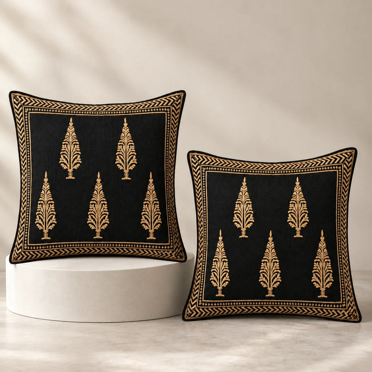 Two square black throw pillows with gold embroidered tree motifs and borders, displayed on a white pedestal against a warm beige background.