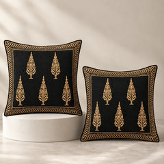Two square black throw pillows with gold embroidered tree motifs and borders, displayed on a white pedestal against a warm beige background.