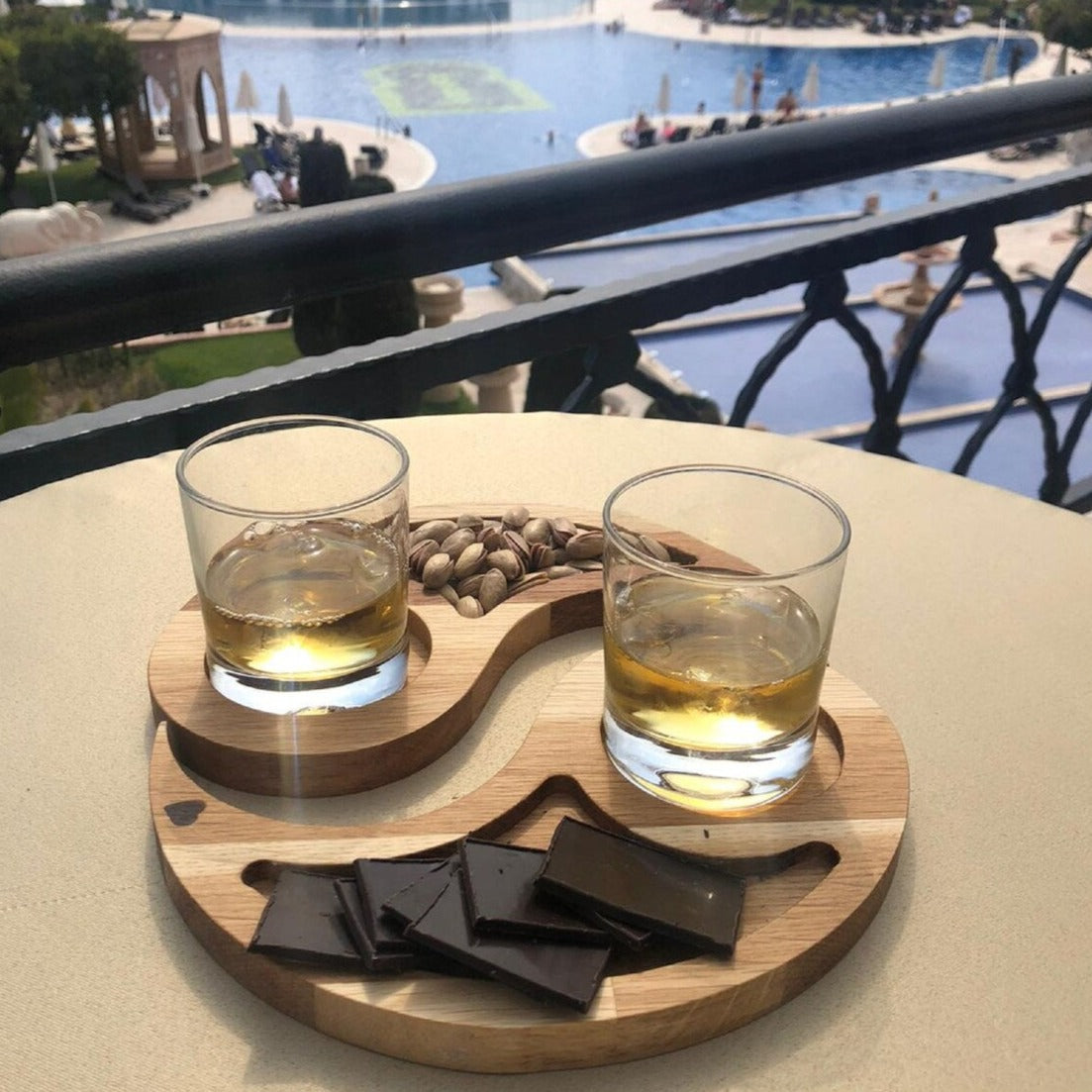 Two curved wood-tone trays holding drinks, nuts and chocolate on a balcony table