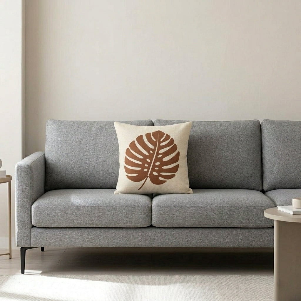 Cream cushion cover with a large terracotta monstera leaf on a grey sofa.