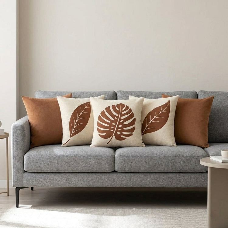 Five cream and terracotta cushion covers with leaf prints and plain designs on a grey sofa.