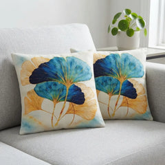 Two cream cushion covers with blue and teal fan-shaped leaves and golden stems on a grey sofa.