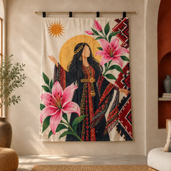 A large wall tapestry featuring a bohemian-style illustration of a woman in a black robe with red and gold patterns, surrounded by large pink lilies, green leaves, and a gold sun on a cream background, hanging on a gold rod in a warm beige room.