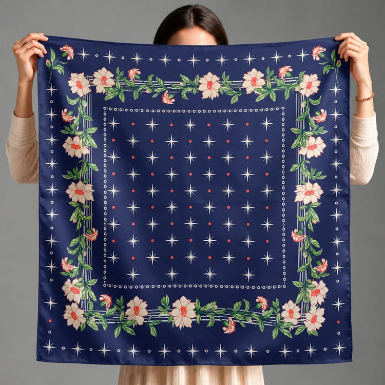A person holds up a square navy blue scarf with a floral border and star pattern against a plain gray background.