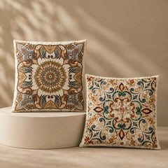 Two decorative throw pillows with ornate bohemian mandala and floral patterns in beige, brown, and teal, displayed on a beige pedestal against a warm beige wall with soft leaf shadows.