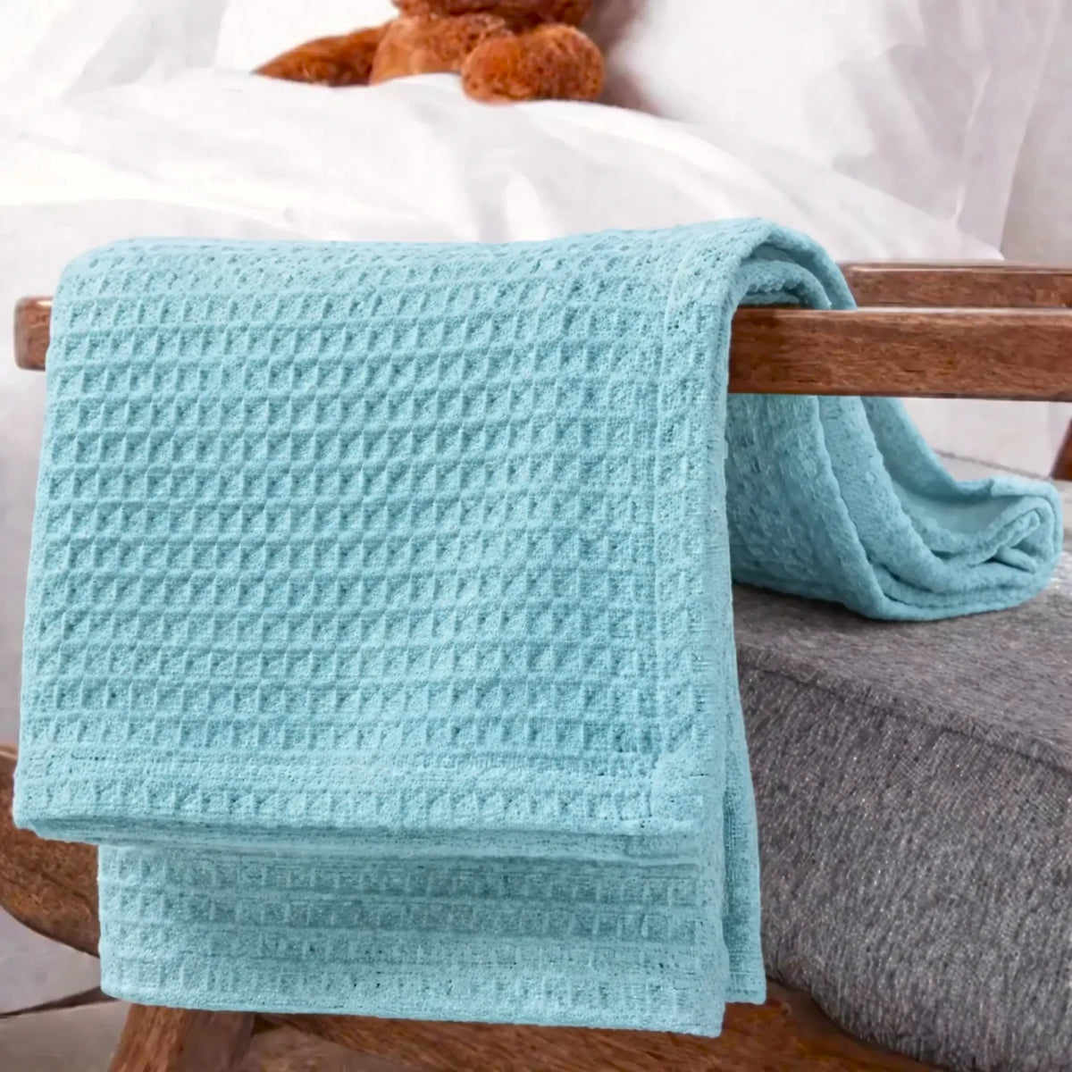 A light aqua blue waffle-weave blanket draped over a wooden chair in a bedroom setting, with a white bed sheet and a brown teddy bear visible in the background.