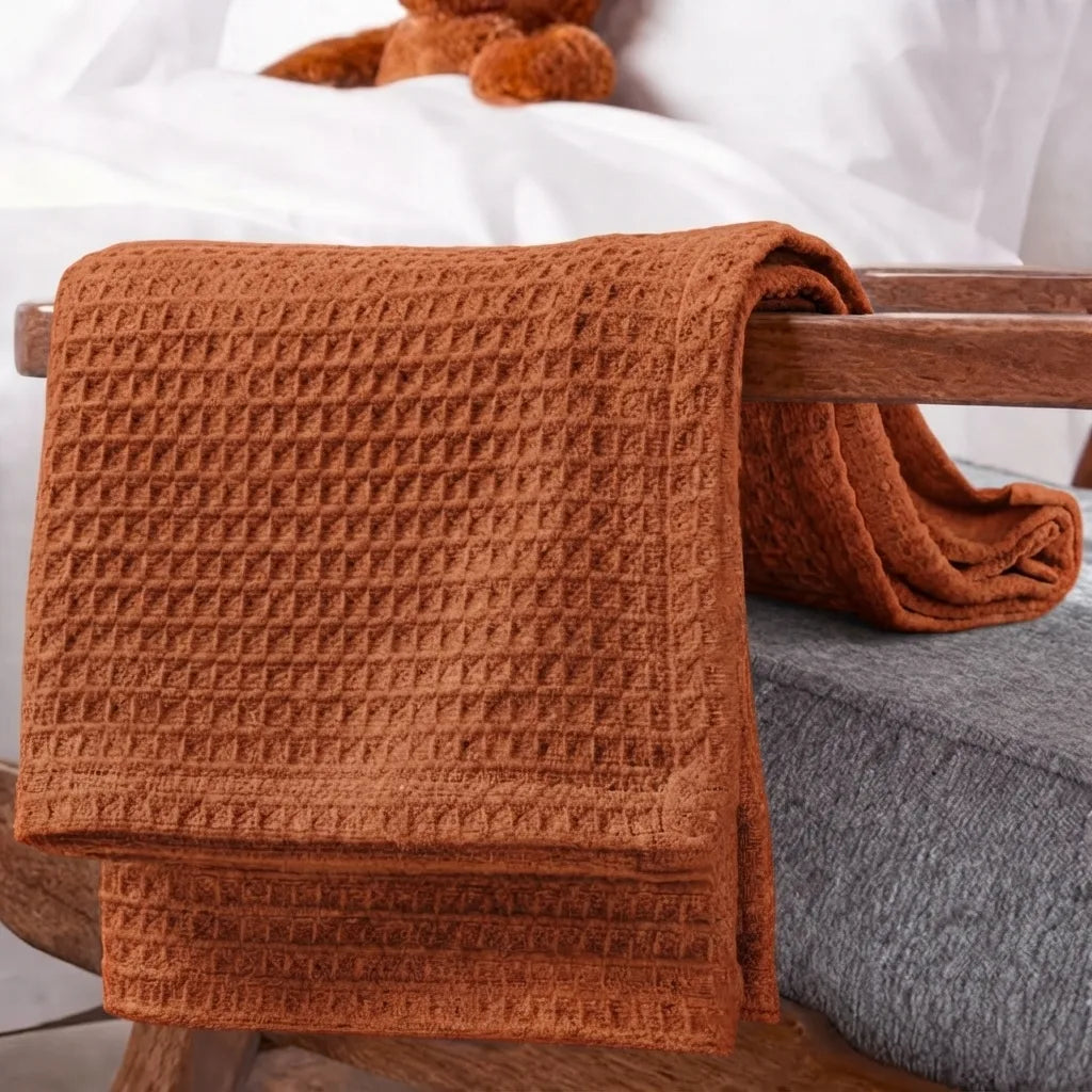 A rust-orange waffle-weave throw blanket draped over a wooden chair in a bedroom setting, with a gray upholstered seat visible underneath and a white bed in the background.