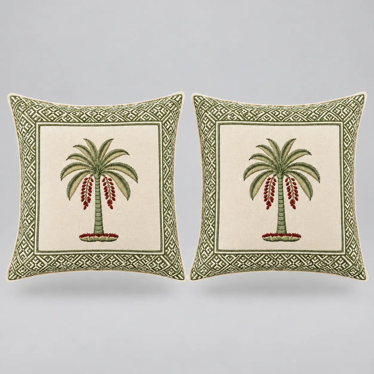 Two square decorative throw pillows shown side by side on a light gray background. Each pillow has an off-white center panel featuring a green palm tree with red hanging fruit, surrounded by a green geometric border.
