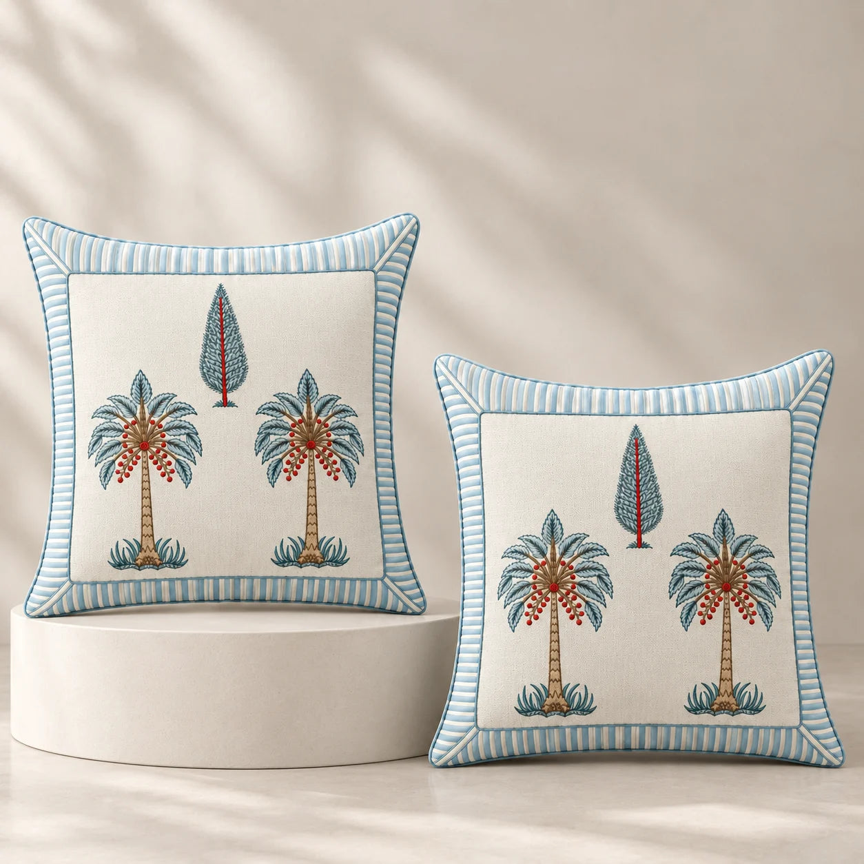 Two decorative square throw pillows with embroidered palm tree motifs and striped blue borders, displayed on a white pedestal against a warm beige background with soft leaf shadows.