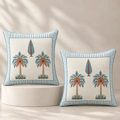 Two decorative square throw pillows with embroidered palm tree motifs and striped blue borders, displayed on a white pedestal against a warm beige background with soft leaf shadows.
