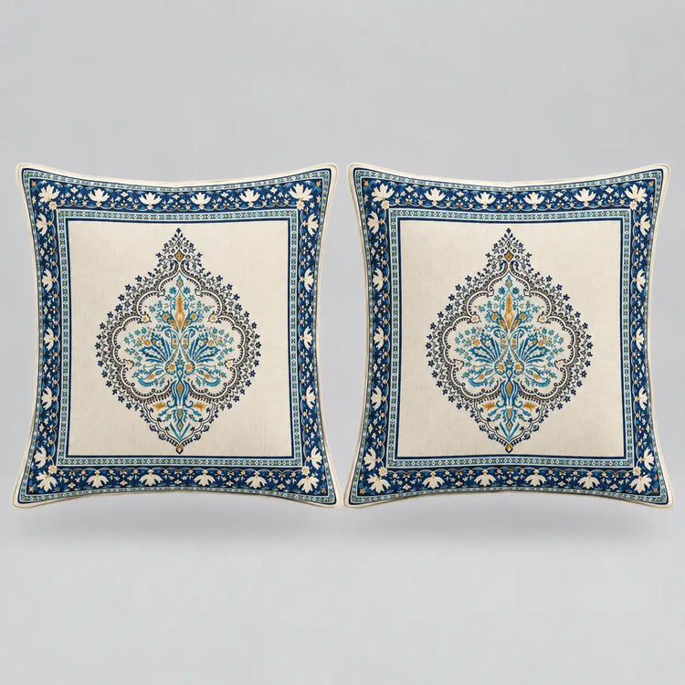 Two square decorative throw pillows with intricate blue and gold traditional patterns on a cream background, set against a neutral gray studio backdrop.