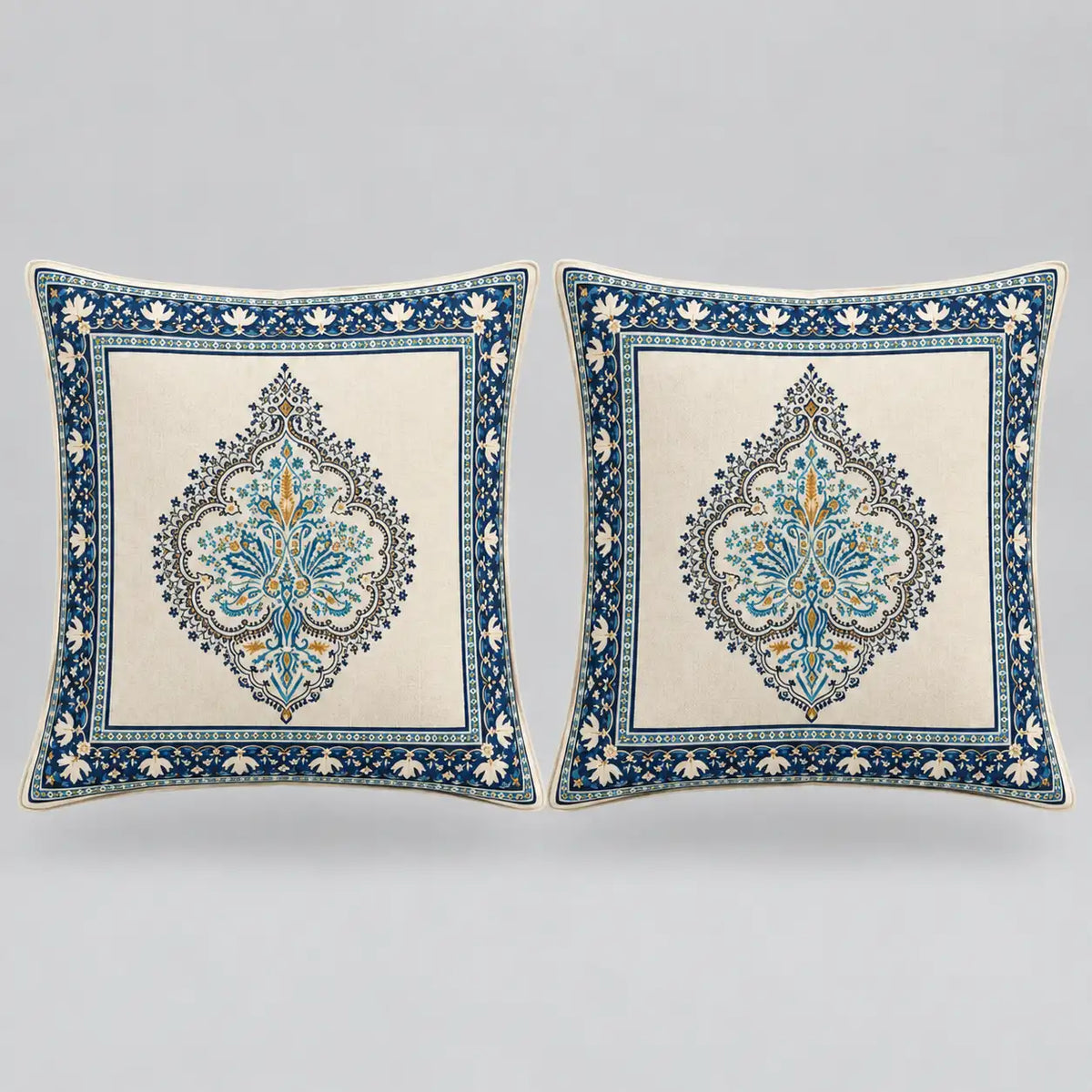Two square decorative throw pillows with intricate blue and gold traditional patterns on a cream background, set against a neutral gray studio backdrop.