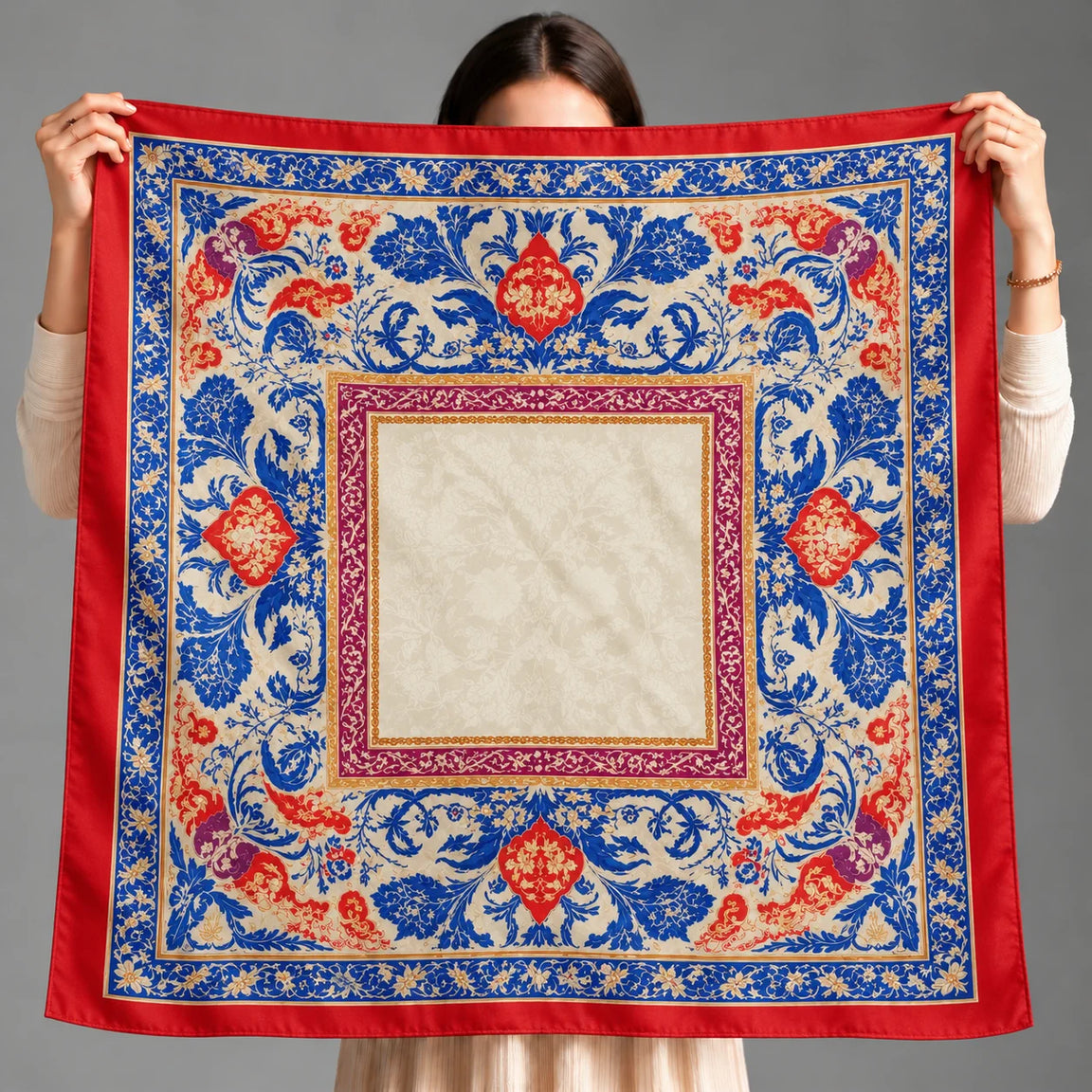 A person holds up a square silk scarf with a traditional Persian-style floral pattern. The scarf features a wide red border, a central cream-colored square, and intricate blue, red, and gold floral motifs.