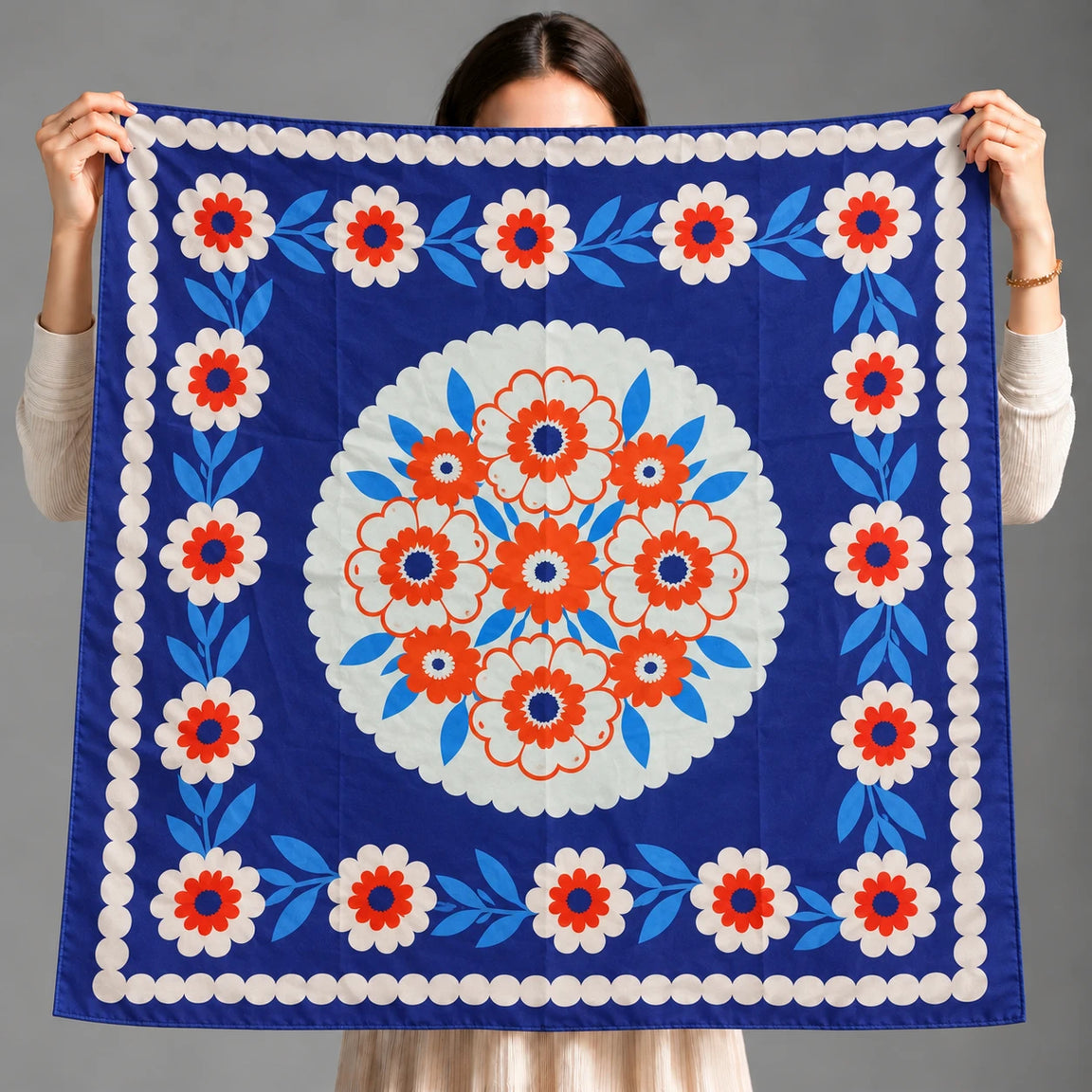 A person holds up a square blue scarf with a white, red, and light blue floral pattern, showing the front design against a plain gray background.