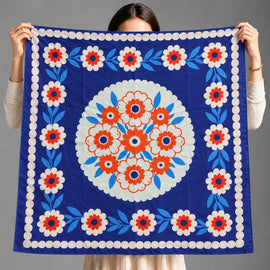A person holds up a square blue scarf with a white, red, and light blue floral pattern, showing the front design against a plain gray background.