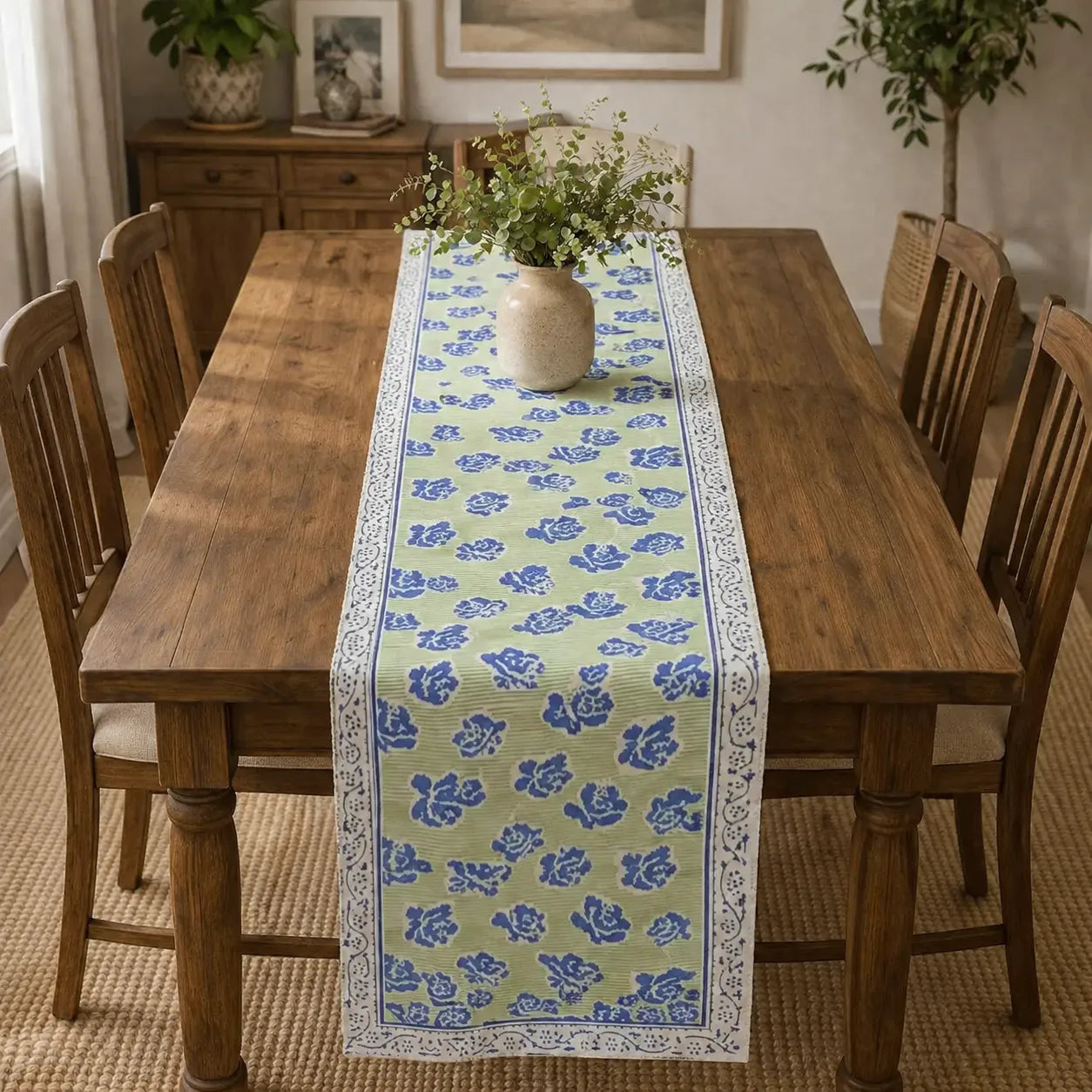 A rectangular wooden dining table in a dining room setting, styled with a light green table runner featuring a blue floral pattern and white borders, topped with a small vase of greenery.