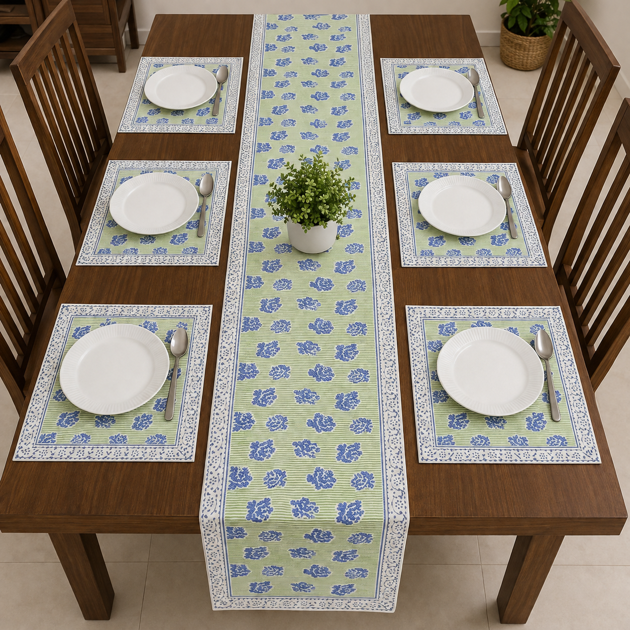 A rectangular wooden dining table set with six matching placemats and a long floral table runner. Each place setting includes a white plate, silver spoon, and folded green napkin, with a small potted green plant centered on the runner.