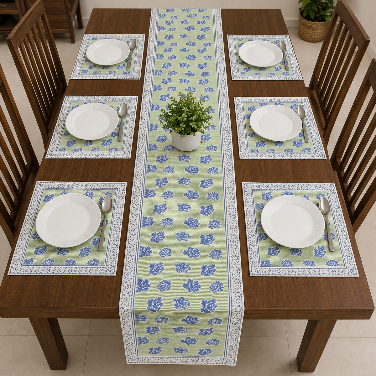 A rectangular wooden dining table set with six matching placemats and a long floral table runner. Each place setting includes a white plate, silver spoon, and folded green napkin, with a small potted green plant centered on the runner.