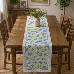 A rectangular wooden dining table in a dining room setting, styled with a light green table runner featuring a blue floral pattern and white borders, topped with a small vase of greenery.