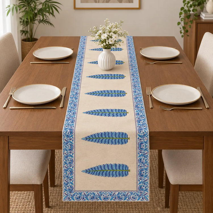 Wooden dining table set with white plates and gold cutlery, featuring a cream-colored table runner adorned with a line of blue and green textured feathers and a blue floral border.   