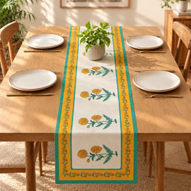 A rectangular table runner with a yellow and green floral pattern is laid down the center of a wooden dining table set with white plates and gold cutlery. A small potted green plant sits on top of the runner.