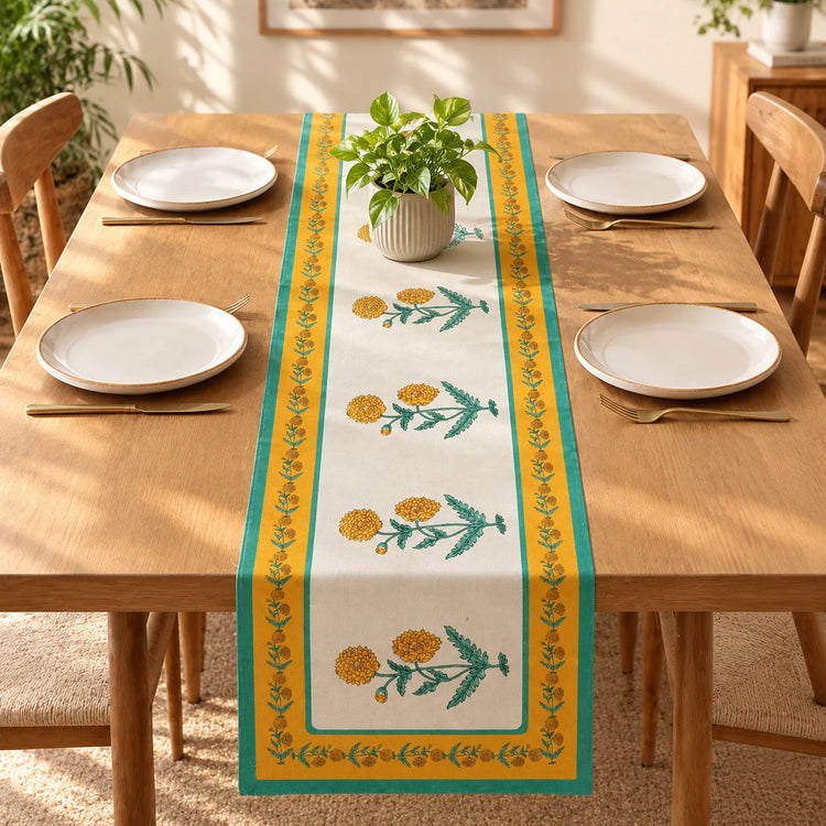 A rectangular table runner with a yellow and green floral pattern is laid down the center of a wooden dining table set with white plates and gold cutlery. A small potted green plant sits on top of the runner.