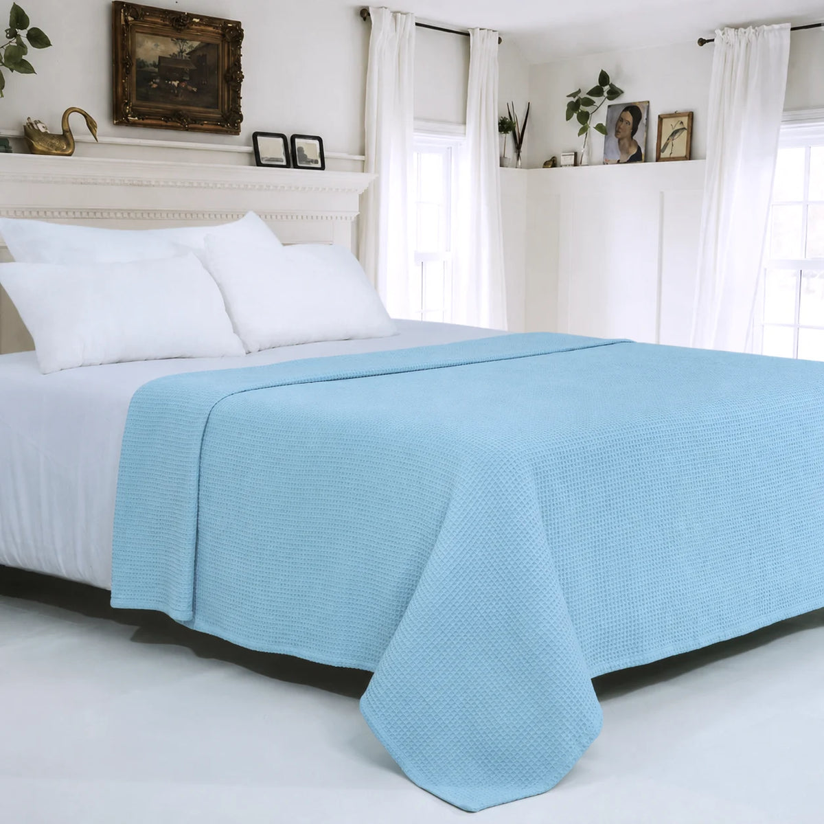 A light blue textured waffle-weave blanket draped over a bed with white sheets and pillows in a bright, white bedroom.