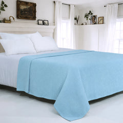 A light blue textured waffle-weave blanket draped over a bed with white sheets and pillows in a bright, white bedroom.