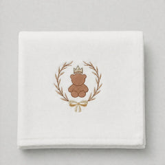 A folded white fabric napkin or handkerchief featuring an embroidered design of a brown teddy bear wearing a small gold crown, surrounded by a laurel wreath and tied with a gold bow.