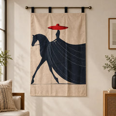 A beige wall hanging or tapestry with a stylized dark navy horse and rider graphic and a bright red hat, suspended from a metal rod in a softly lit room.