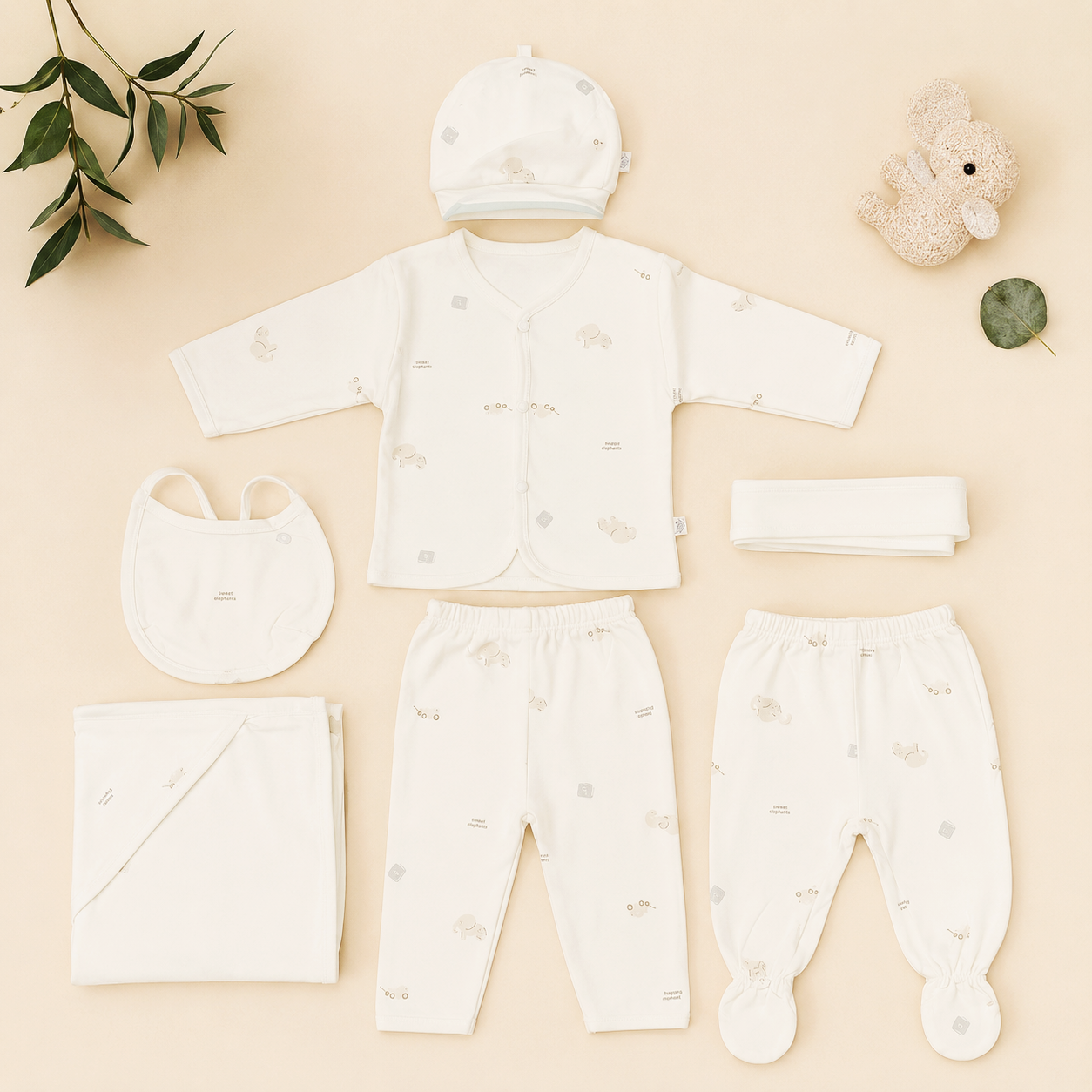A baby clothing set arranged on a light beige background, including a cream long-sleeve top, matching pants, a hat, a bib, a folded blanket, a folded cloth, and a folded headband, with small animal prints. Decorative props include a plush bunny, a leaf, and a folded leaf in the corners.