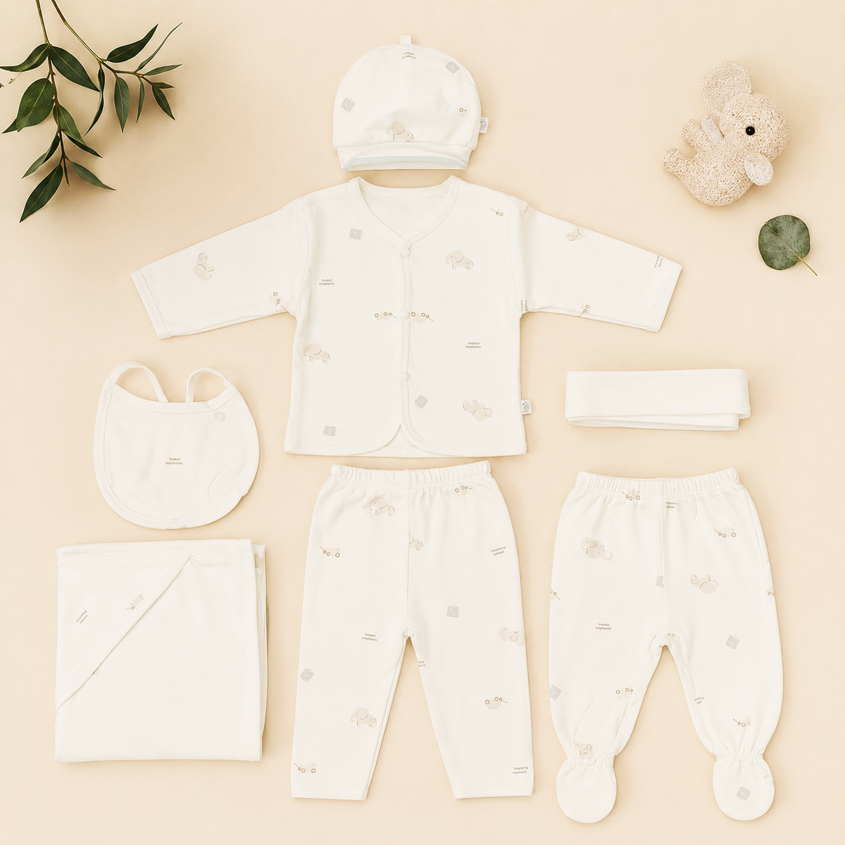 A baby clothing set arranged on a light beige background, including a cream long-sleeve top, matching pants, a hat, a bib, a folded blanket, a folded cloth, and a folded headband, with small animal prints. Decorative props include a plush bunny, a leaf, and a folded leaf in the corners.