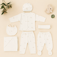 A baby clothing set arranged on a light beige background, including a cream long-sleeve top, matching pants, a hat, a bib, a folded blanket, a folded cloth, and a folded headband, with small animal prints. Decorative props include a plush bunny, a leaf, and a folded leaf in the corners.