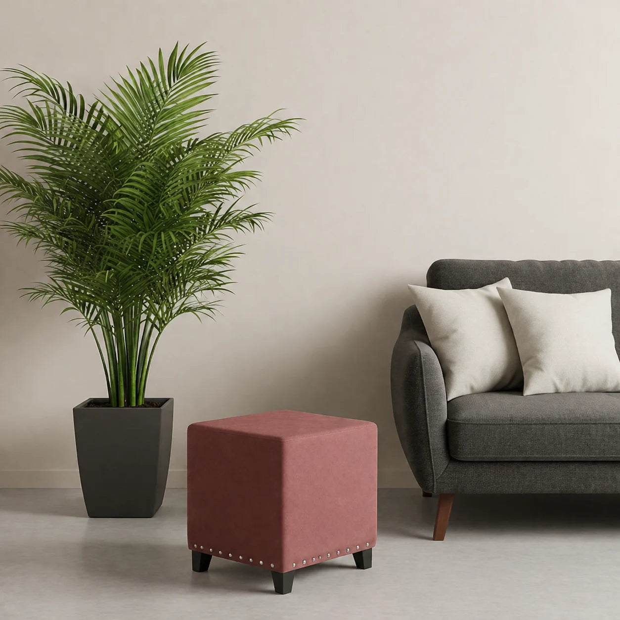 A dusty rose pink upholstered cube ottoman with dark legs and silver nailhead trim, styled in a living room next to a potted palm and a gray sofa.