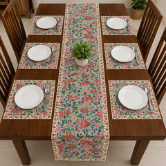 A floral table runner and matching placemats arranged on a dark wood dining table set with white plates, silver cutlery, and wooden chairs. A small potted green plant sits in the center of the table.