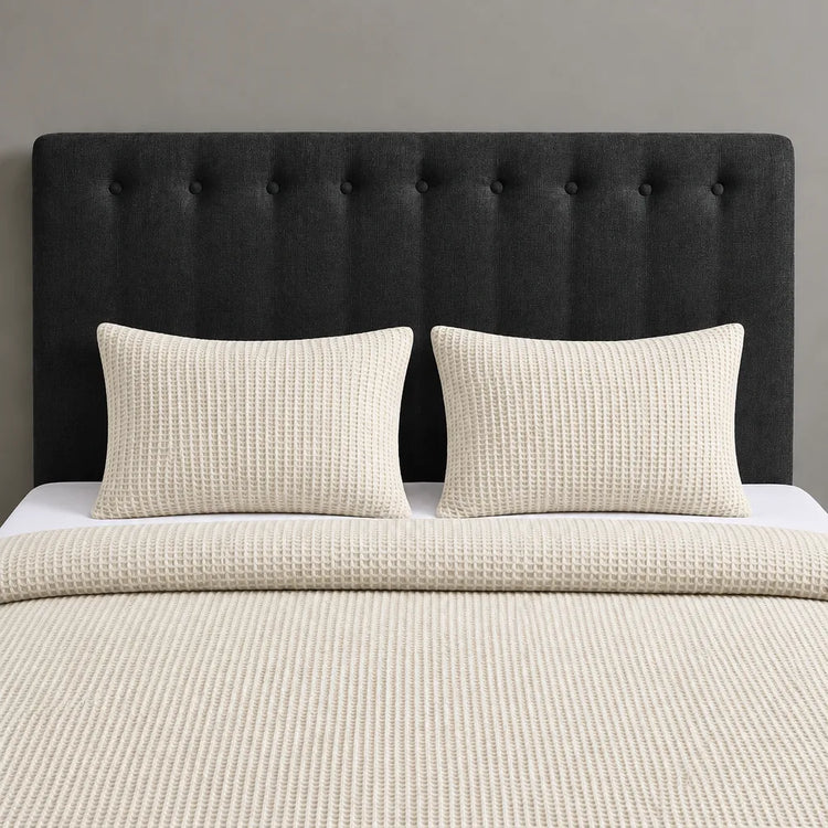 A neatly made bed with a textured cream waffle blanket, two matching cream pillows, and a tall black tufted headboard against a gray wall.