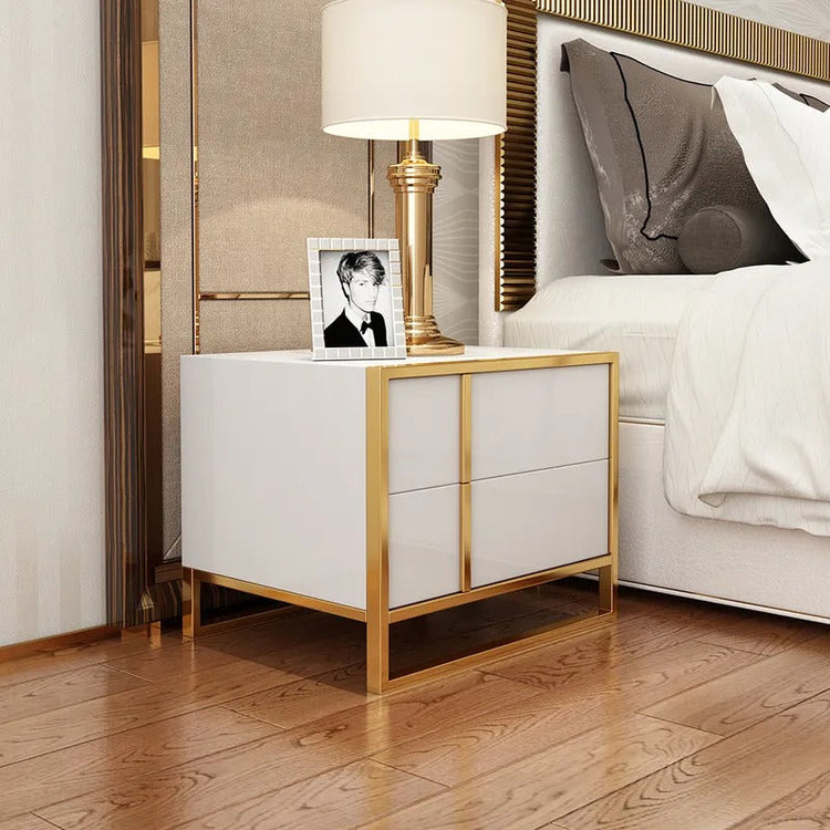 Angled view of a white bedside cabinet with gold-colored frame beside a bed.
