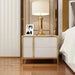 White two-drawer bedside cabinet with gold-colored trim holding a lamp and framed photograph.
