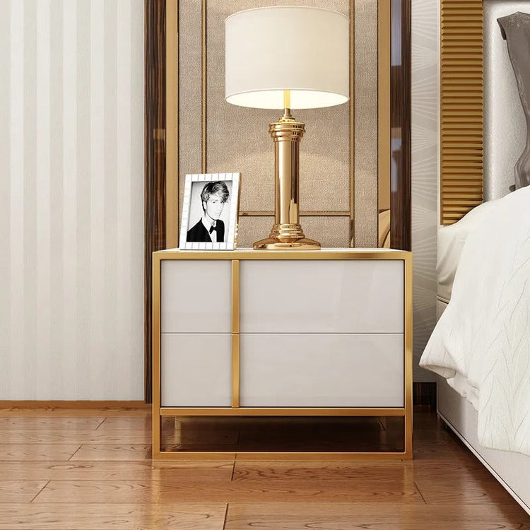 White two-drawer bedside cabinet with gold-colored trim holding a lamp and framed photograph.