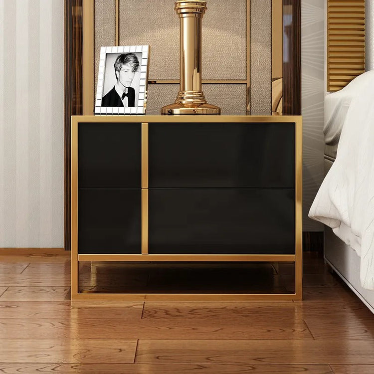 Black two-drawer bedside cabinet with gold-colored trim holding a lamp and framed photograph.