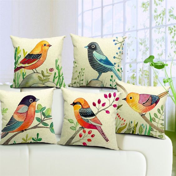 Five cream cushion covers with colourful bird illustrations arranged on a sofa.