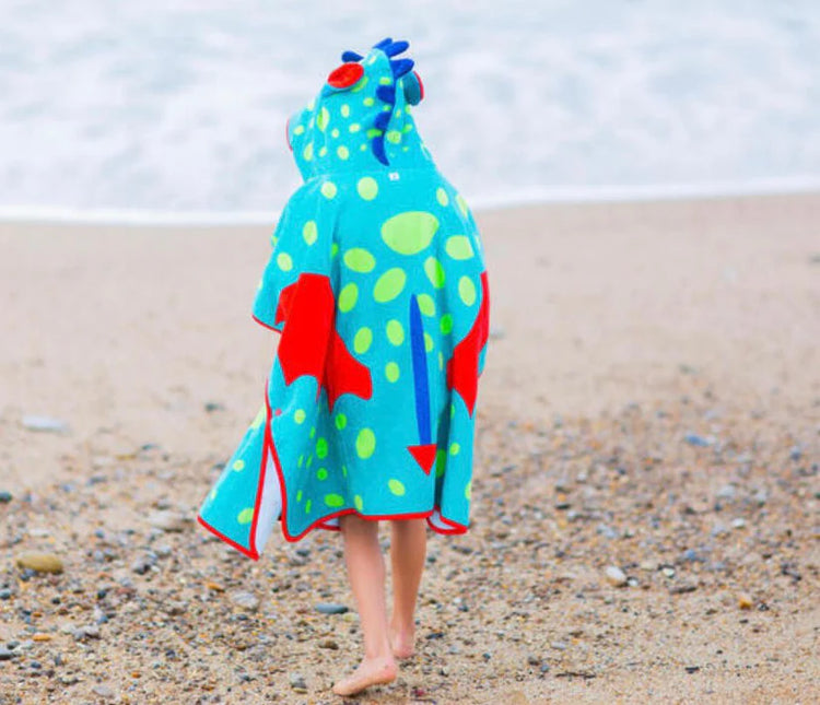 Back view of a child wearing a turquoise dinosaur towel poncho on a beach.