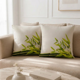 Two square off-white throw pillows with a green bamboo leaf print, styled on a beige fabric sofa in a bright, modern living room.