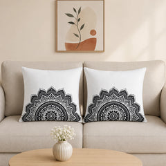 Two square white throw pillows with black mandala patterns, styled on a beige sofa in a living room setting with a small table and vase in the foreground and framed wall art above.