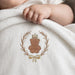 A close-up of a baby's hand resting on a white textured blanket featuring an embroidered brown teddy bear wearing a small gold crown, surrounded by a laurel wreath and tied with a bow.