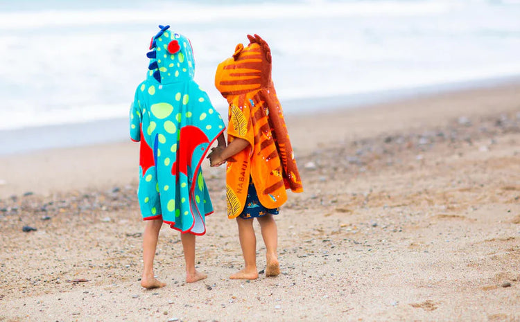 Two children wearing turquoise dinosaur and orange animal ponchos at the beach.
