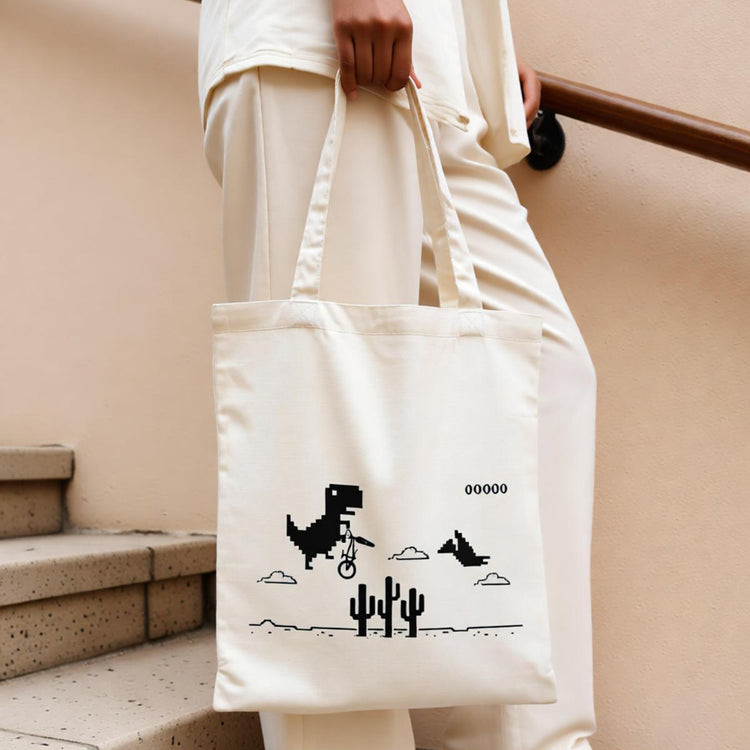 Cream tote bag with a pixel-style dinosaur and cactus print.