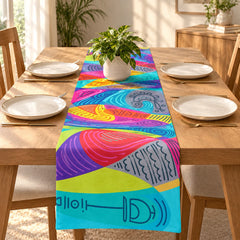 A colorful abstract-patterned table runner laid down the center of a wooden dining table set with white plates, gold cutlery, and a potted green plant in a bright, sunlit room.