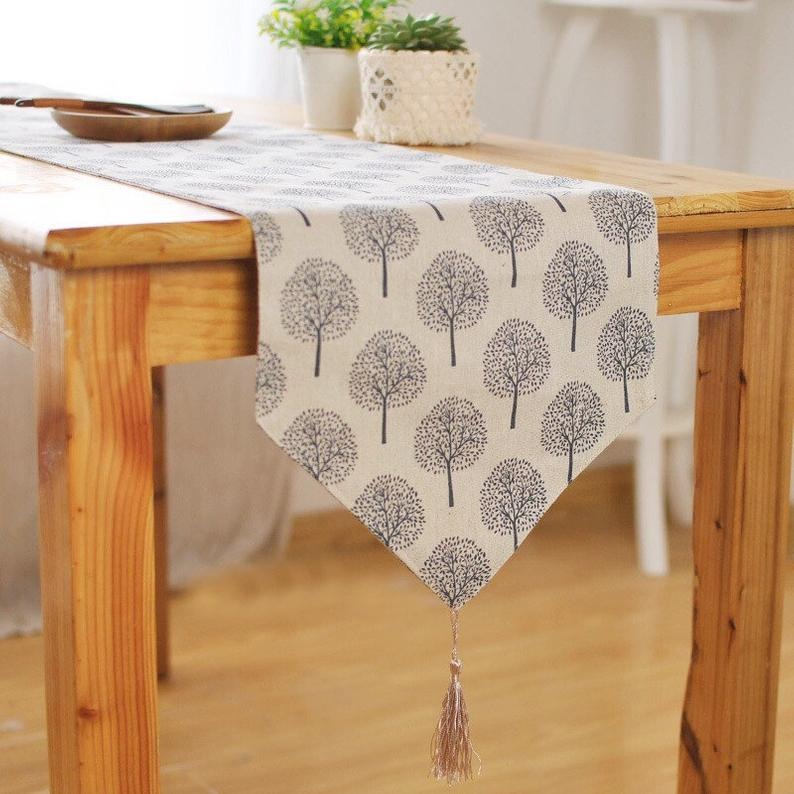 Light beige table runner with grey tree motifs and a tasselled end.