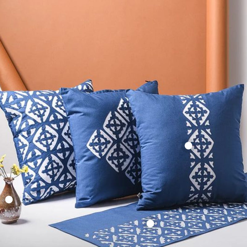A set of three square blue cushion covers with a geometric flower pattern, displayed on a sofa with a tan background. The covers are adorned with a vibrant floral design in a geometric style.